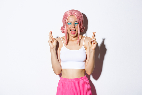Image of optimistic party girl in pink wig, with halloween makeup, close eyes and screaming excited while cross fingers to make a wish, standing over white background.