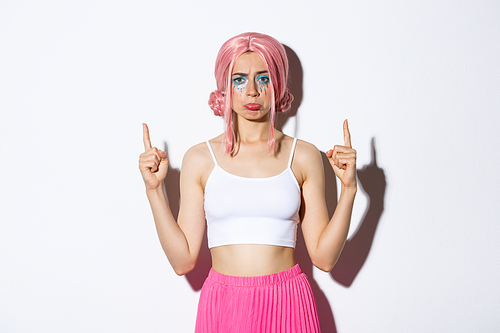 Portrait of disappointed silly girl in pink wig, pointing fingers up and sulking upset, complaining about something bad, standing over white background.