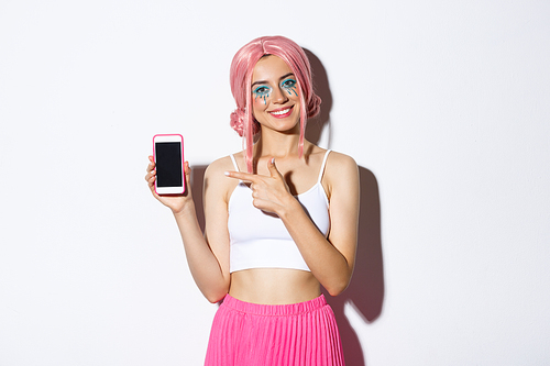 Portrait of happy beautiful female model in pink glamour wig and bright makeup, pointing finger at mobile phone screen, showing application or banner.