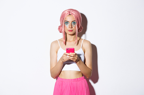 Portrait of attractive party girl in pink wig, with bright makeup, holding smartphone and looking at camera confident.