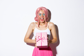 Cheerful birthday girl looking excited, wearing pink wig, shouting of joy, receiving bday gift, standing over white background and celebrating.