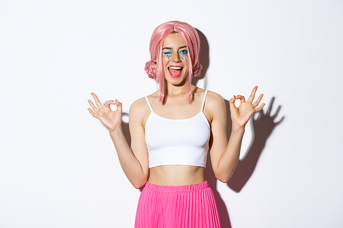Cheerful party girl with bright makeup and pink wig recommend something, showing okay signs and winking satisfied, standing in halloween costume over white background.