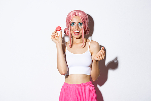 Image of beautiful girl with bright makeup eating tasty macaroons, wearing pink wig, showing tongue and smiling, standing over white background.