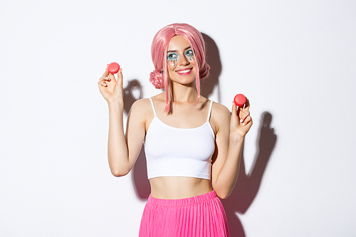 Lovely young woman in pink wig showing macaroons and smiling, eating sweets, standing over white background.