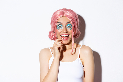 Close-up of excited party girl in pink wig and bright makeup looking impressed, smiling and gazing surprised, standing over white background.