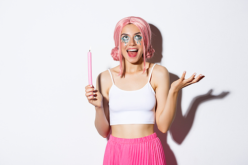 Portrait of excited beautiful girl dressed up as fairy on halloween, wearing pink wig and bright makeup, holding candle and talking to someone, standing over white background.