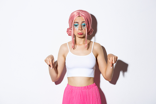 Portrait of gloomy cute girl in pink wig and party outfit, looking disappointed and pouting, pointing fingers down at bad news, standing over white background.