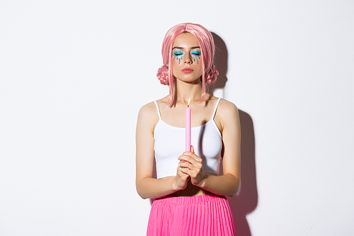 Image of pretty girl in pink wig, with bright makeup, dressed up as fairy for halloween party, close eyes while holding candle and casting magic spell, standing over white background.