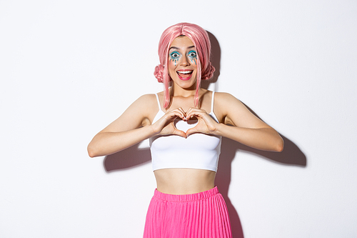 Image of excited smiling girl like something, showing heart gesture and looking amazed, wearing halloween costume with bright makeup and pink wig.