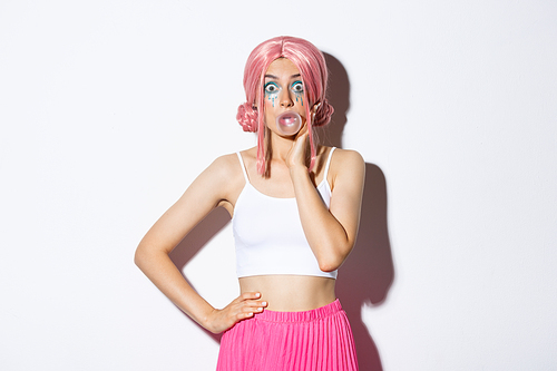 Portrait of surprised pretty girl in pink wig, blowing bubble gum and looking amazed at camera, standing over white background.