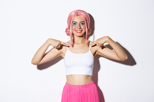 Portrait of cheerful party girl with pink wig and bright makeup, pointing at herself and smiling confident, showing her halloween outfit, standing over white background.