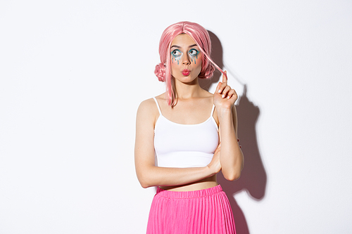 Silly young party girl in pink wig and halloween makeup, looking at upper left corner thoughtful, standing coquettish over white background.