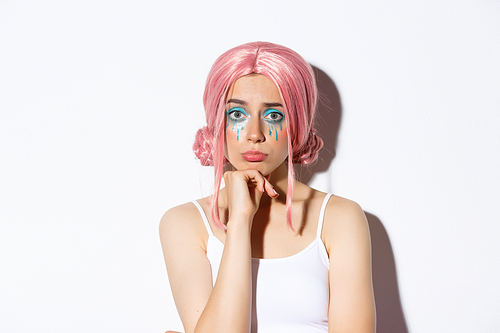 Close-up of sad cute girl with halloween makeup and pink wig looking upset, sulking while standing over white background.