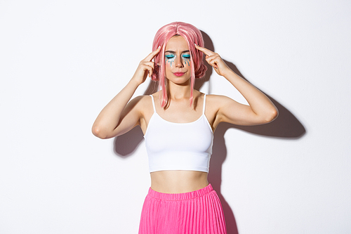 Portrait of distressed caucasian girl with pink wig and party makeup, standing with closed eyes and touching head, trying to think or remember something.