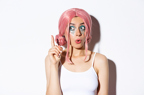 Close-up of woman in pink party wig and bright makeup, have idea, raising index finger in eureka sign, standing over white background.