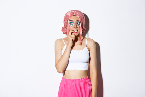 Image of silly beautiful girl with pink wig and halloween makeup, biting finger and looking tempted left, want something, standing over white background.