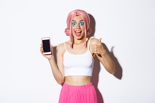 Excited stylish girl in pink wig, showing thumbs-up and looking fascnated while showing something awesome on mobile phone screen, standing over white background.