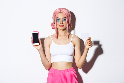 Image of stylish party girl in pink wig, showing credit card and mobile phone screen, celebrating holiday in glamour outfit, standing over white background.