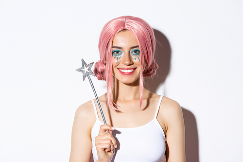 Close-up of beautiful girl dressed-up as a fairy for halloween party, holding magic wand and smiling, standing over white background.