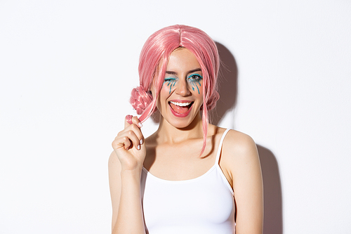 Portrait of sassy attractive girl in pink wig, wearing outfit for halloween party, smiling and winking at camera, standing over white background.