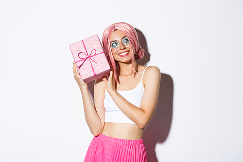 Image of beautiful girl in pink wig shaking box with birthday gift, wonder what inside wrapped box, standing over white background.