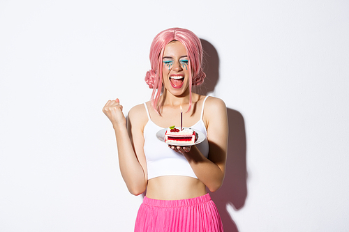 Portrait of cheerful smiling girl celebrating her birthday, wearing pink wig, holding b-day cake and shouting of joy, standing over white background.