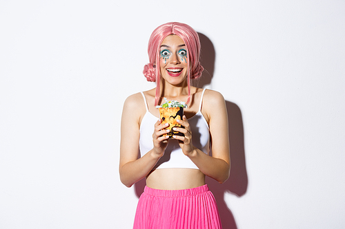 Portrait of excited girl celebrating halloween, enjoy trick or treat, holding candies and smiling happy at camera, dressed in pink anime wig, standing over white background.