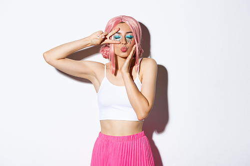 Image of attractive glamour girl in pink wig, bright halloween makeup, standing over white background with peace sign and closed eyes, kissing someone.