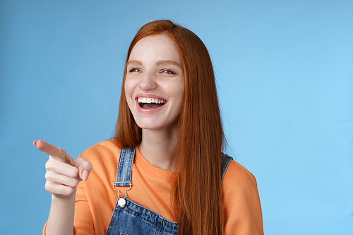 Adorable fascinated young attractive tender redhead amused girl having fun pointing looking left astonished laughing having fun see funny advertisement chuckling joyfully, blue background.