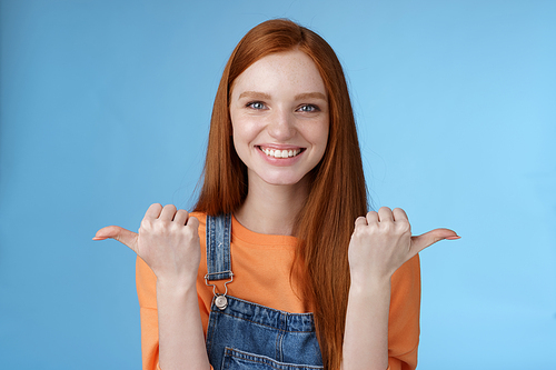 Indoor shot charismatic assertive happy smiling redhead woman orange shirt denim overalls pointing sideways thumbs left right showing choices opportunities give chance choose, blue background.