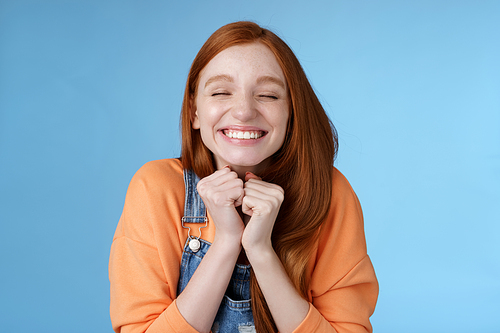 Eager rejoicing thrilled pretty young redhead girl close eyes dreamy smiling receive great result scholarchip triumphing joyfully grinning squeez hands excited, standing blue background very happy.