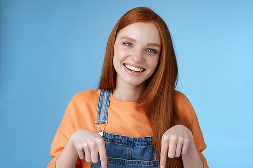 Friendly happy redhead girl smiling lively pretty grin pointing down index fingers offering good offer recommend use service standing blue background discuss interesting product, blue background.