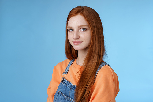Lifestyle. Waist-up kind sincere tender lovely redhead girl wearing orange shirt denim overalls standing half-turned smiling silly gentle grin camera looking friendly pleasantly walk alone blue background.