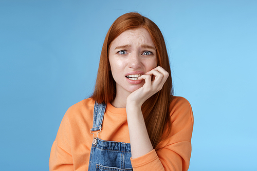 Intense worried scared unsure young redhead panicking silly girl frowning look upset anxious biting fingernails emotional frightened fired standing blue background emotional terrified. Copy space