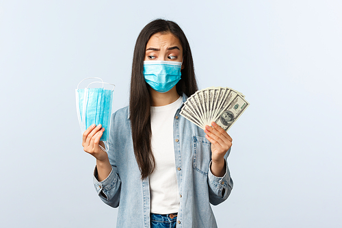 covid-19 pandemic, coronavirus expences and finance concept. Thoughtful and puzzled asian female looking at medical mask and showing money, losing money during coronavirus.