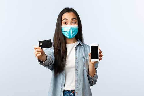 Social distancing lifestyle, covid-19 pandemic and contactless shopping concept. Cheerful pretty asian girl in medical mask advice using credit card for paying online for orders.