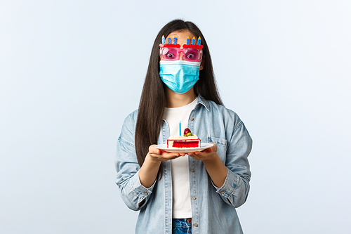 Social distancing lifestyle, covid-19 pandemic, celebrating holidays during coronavirus concept. Cheerful birthday girl in medical mask and party glasses, holding b-day cake, enjoying celebration.