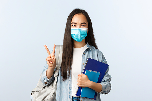 Covid-19 pandemic, education during coronavirus, back to school concept. Cute asian college student in medical mask, carry notebooks and backpack, show peace sign, starting new semester.