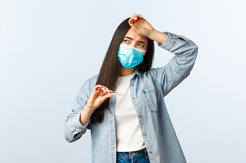 Social distancing lifestyle, covid-19 pandemic everyday life and leisure concept. Sick woman with coronavirus disease, having high fever, touch forehead and showing thermometer, wear medical mask.