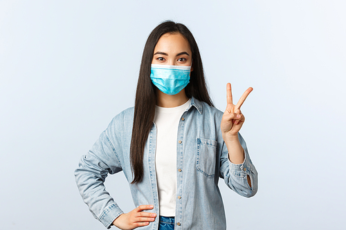 Social distancing lifestyle, covid-19 pandemic and people emotions concept. Cute asian girl in medical mask making order, showing two fingers or peace sign, stand white background.