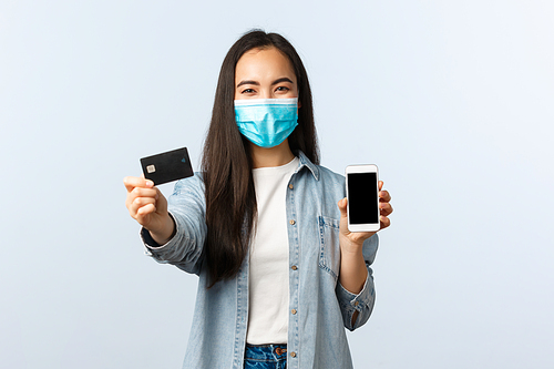 Social distancing lifestyle, covid-19 pandemic and contactless shopping concept. Cheerful friend asian girl in medical mask showing credit card and mobile screen, advice order online and pay.