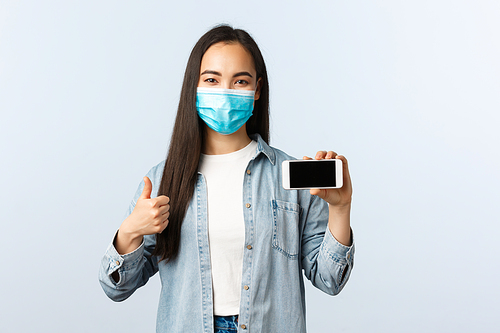 Social distancing lifestyle, covid-19 pandemic and technology concept. Satisfied korean girl in medical mask, thumbs-up while recommend smartphone app, mobile phone store.