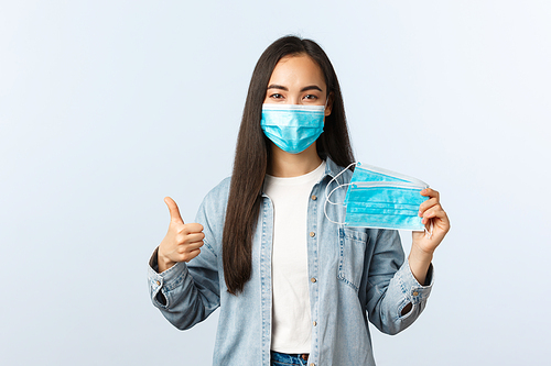 Social distancing lifestyle, covid-19 pandemic everyday life and leisure concept. Cute friendly asian girl showing medical mask and thumbs-up, recommend protect health from coronavirus disease.