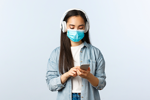 Social distancing lifestyle, covid-19 pandemic and self-isolation leisure concept. Modern cute asian girl in medical mask, listening music wireless headphones, using mobile phone app.