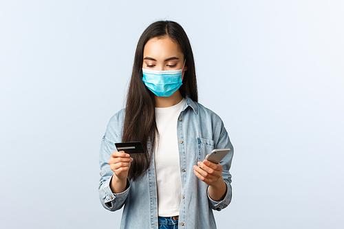 Social distancing lifestyle, covid-19 pandemic and contactless shopping concept. Attractive stylish asian girl in medical mask looking at credit card while making order on mobile application.