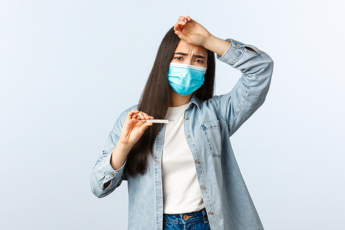 Social distancing lifestyle, covid-19 pandemic everyday life and leisure concept. Sick woman with coronavirus disease, having high fever, touch forehead and showing thermometer, wear medical mask.