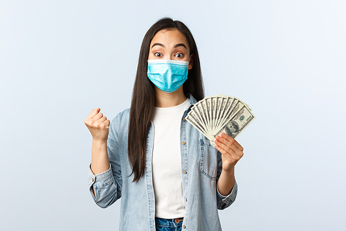 Social distancing lifestyle, covid-19 pandemic business and employement concept. Excited happy asian woman winning lottery, wear medical mask and rejoicing, show money, holding cash.