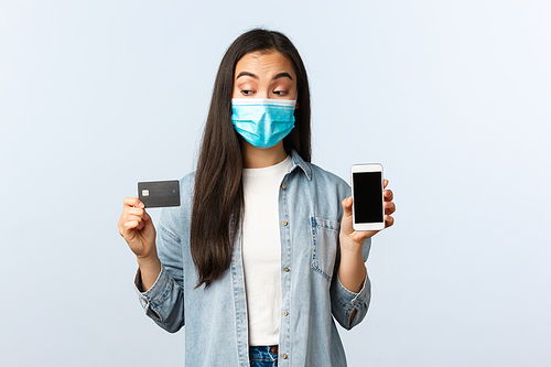 Social distancing lifestyle, covid-19 pandemic and contactless shopping concept. Intrigued cute asian girl in medical mask peeking at credit card, showing smartphone display.
