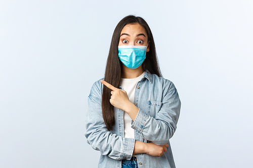 Social distancing lifestyle, covid-19 pandemic everyday life and leisure concept. Excited good-looking asian girl in medical mask talking about new product in store, pointing finger left.
