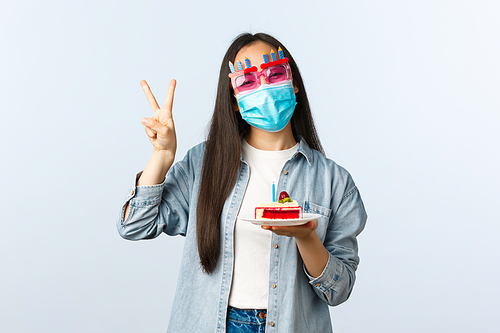 Social distancing lifestyle, covid-19 pandemic, celebrate holidays on coronavirus concept. Happy smiling asian b-day girl in medical mask and party glasses, holding birthday cake and show peace sign.